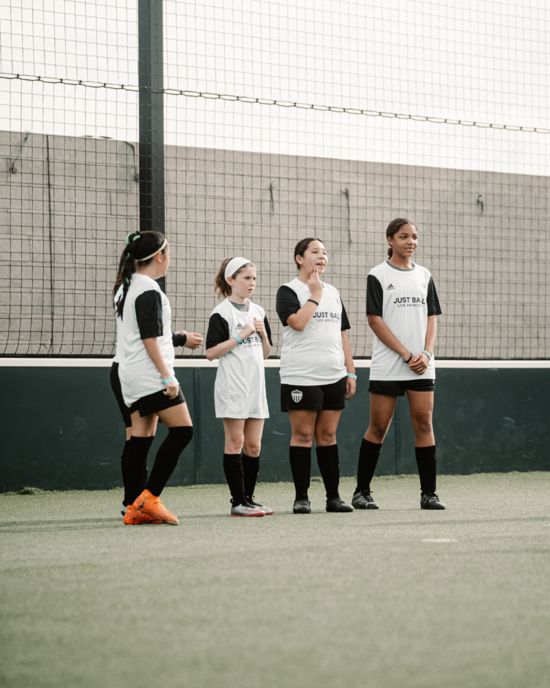 A group of four girls stand and talk on a soccer pitch.
